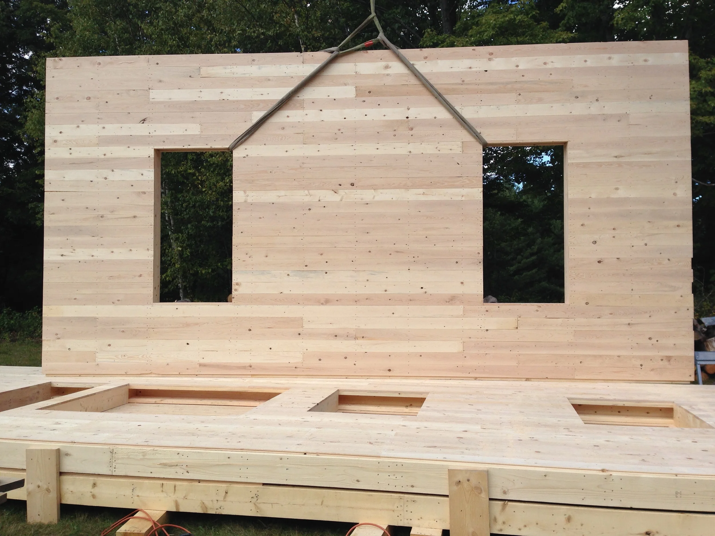 Wooden wall frame of a house under construction with two windows and a partially assembled floor, outdoors with trees in the background. Construction by Corrigan & Klein Artisan Builders.