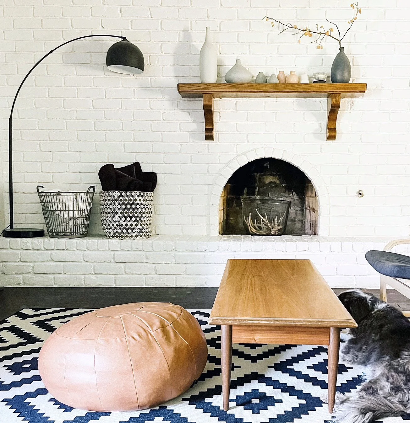 Living room with white brick wall, wooden coffee table, black and white patterned rug, tan leather pouf, black and white baskets, black floor lamp, decorative vases on wooden shelf, and a dog lying on the floor