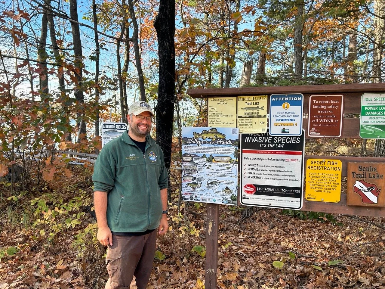 Mark Luhering is standing near the infographic which is on a wooden sign post next to other fishing regulations. There is a lake and fall leaves in the background.