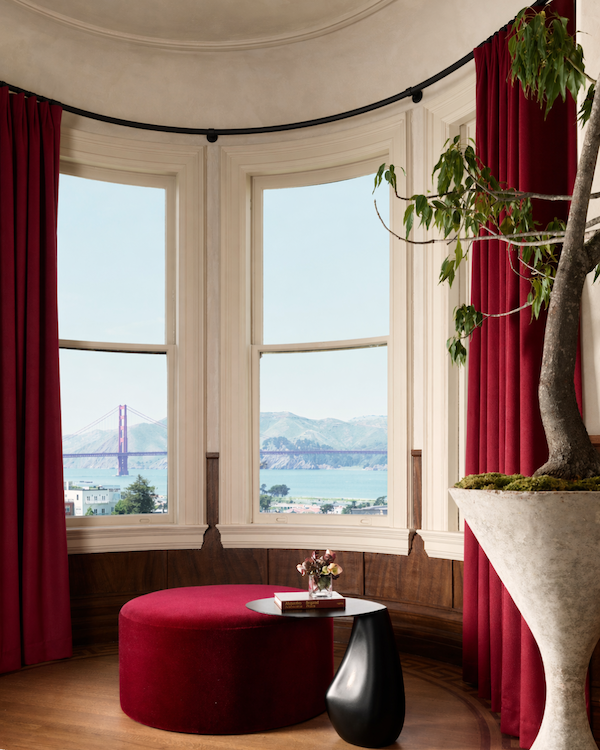 Round ottoman reupholstered by Revitaliste in Dedar Milano Vladimiro mohair in bordeaux, positioned in the curved bay window of Castellanos Interiors' West Red bedroom at the 2026 San Francisco Decorator Showcase, with views of the Golden Gate Bridge