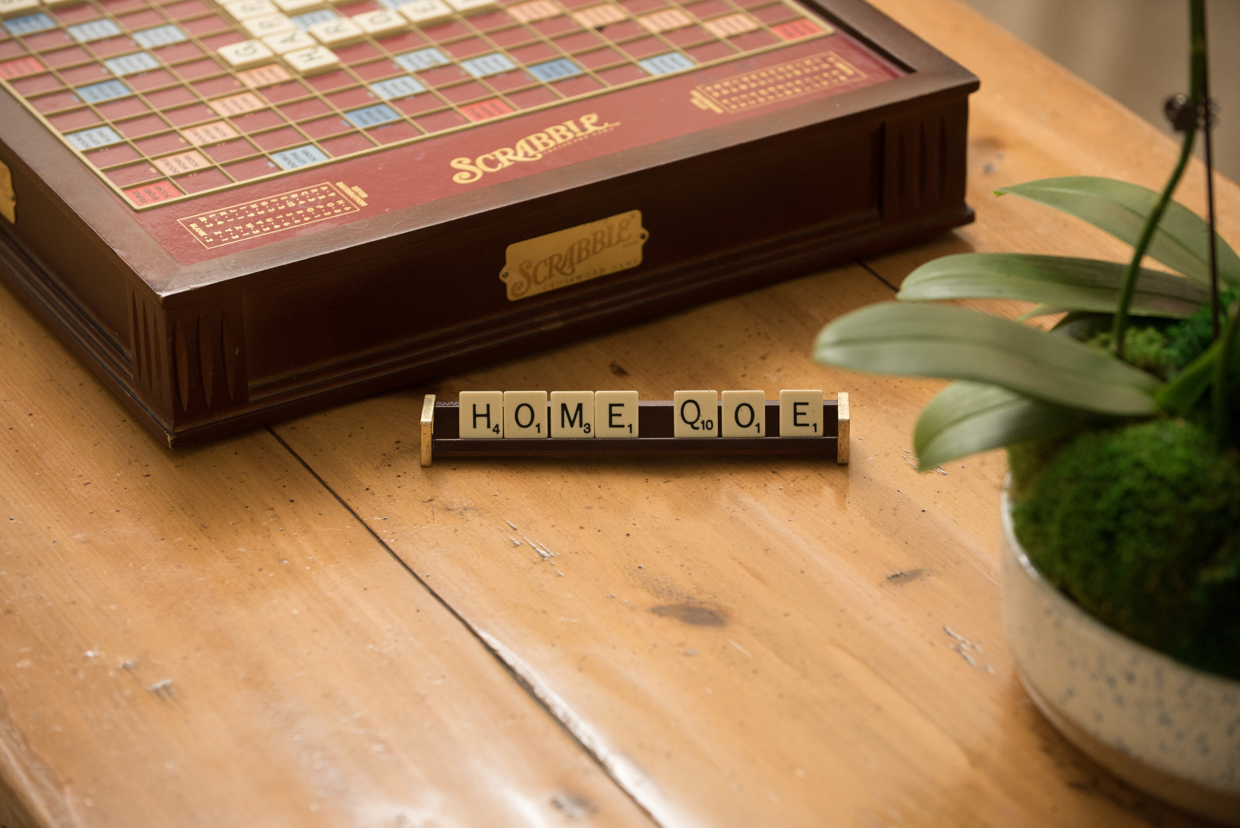 Scrabble game, tile holder with tiles spelling "home" on brown table next to green plant.