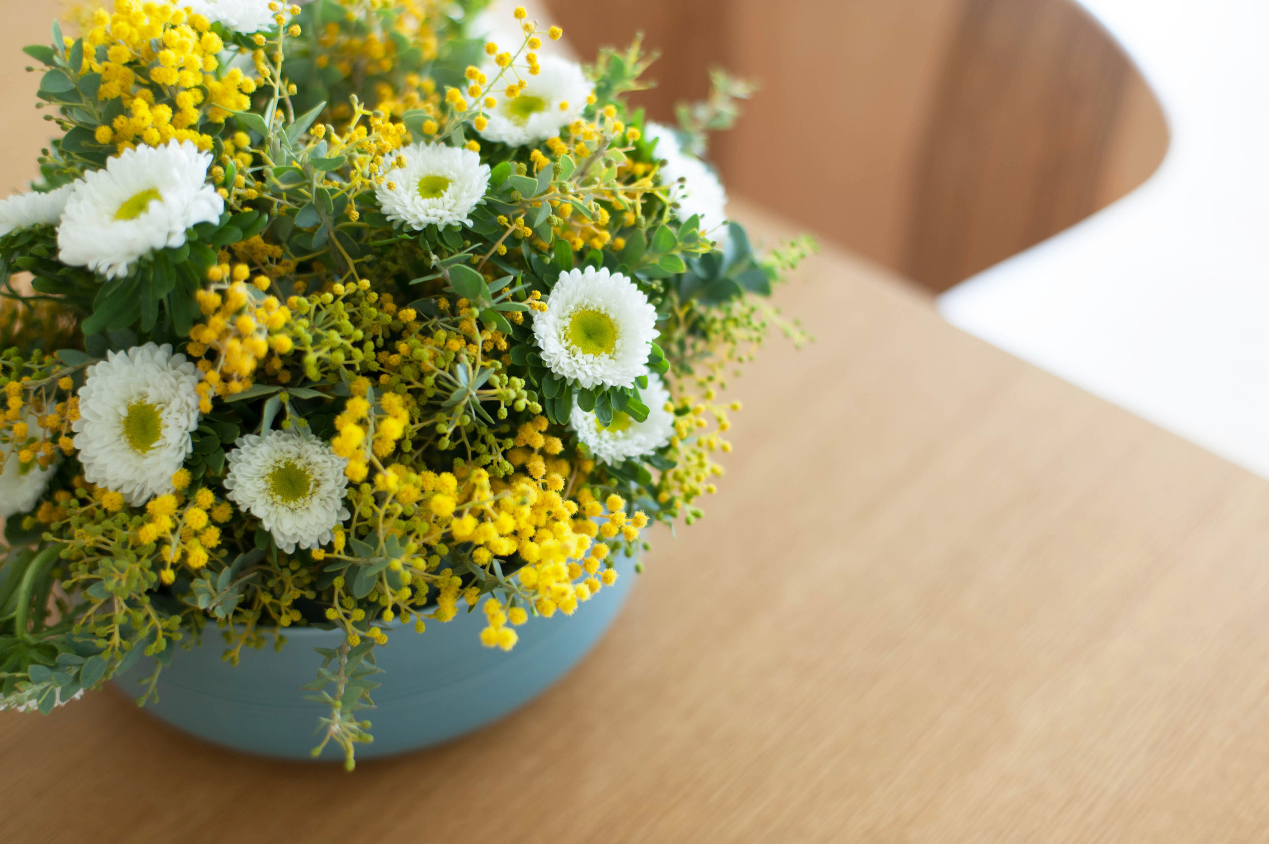 Yellow white and green floral arrangement in blue planter on wooden table