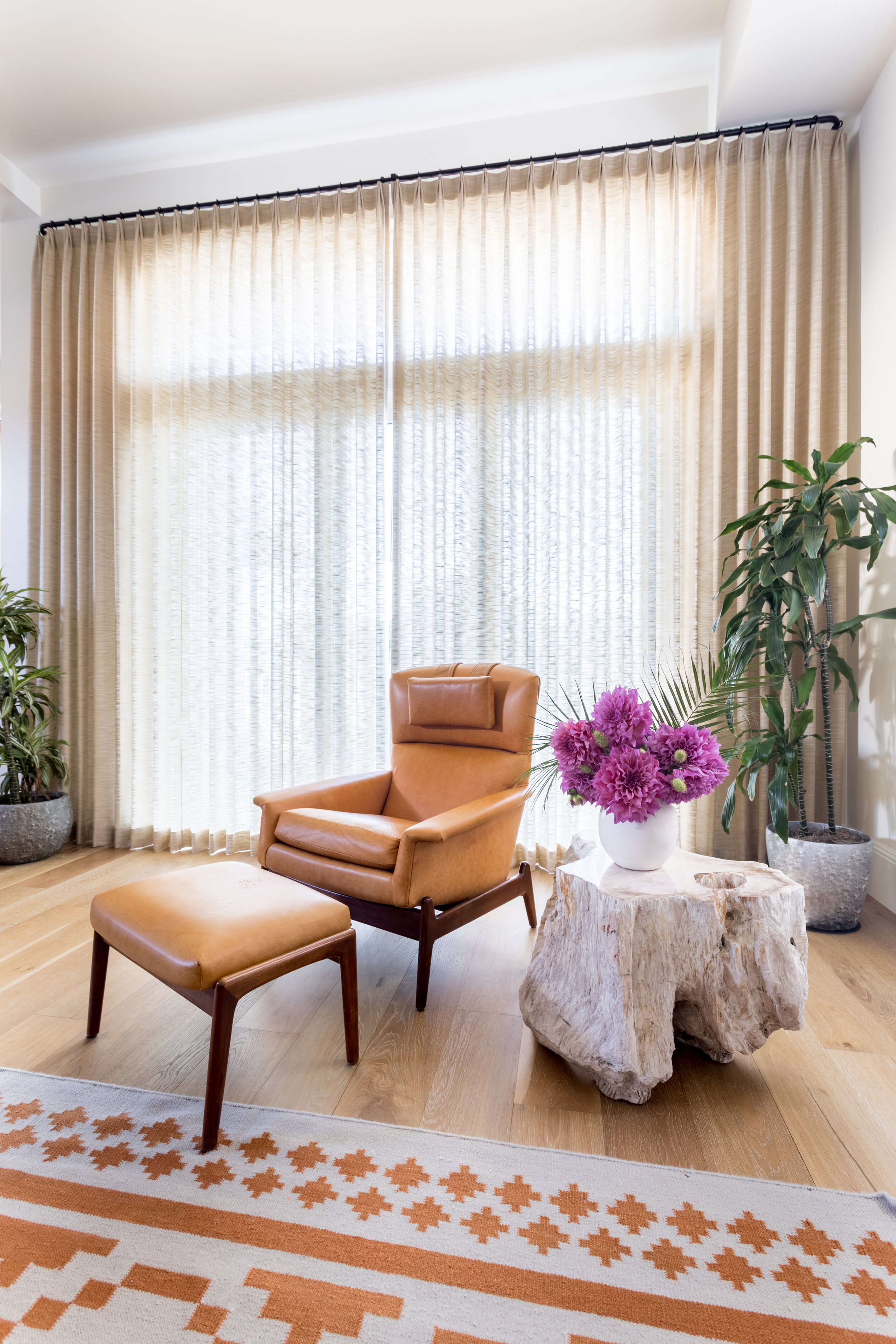 Orange leather chair and ottoman with brown wooden legs next to wooden side table with white vase and purple flowers. Green plants and beige drapery in background.