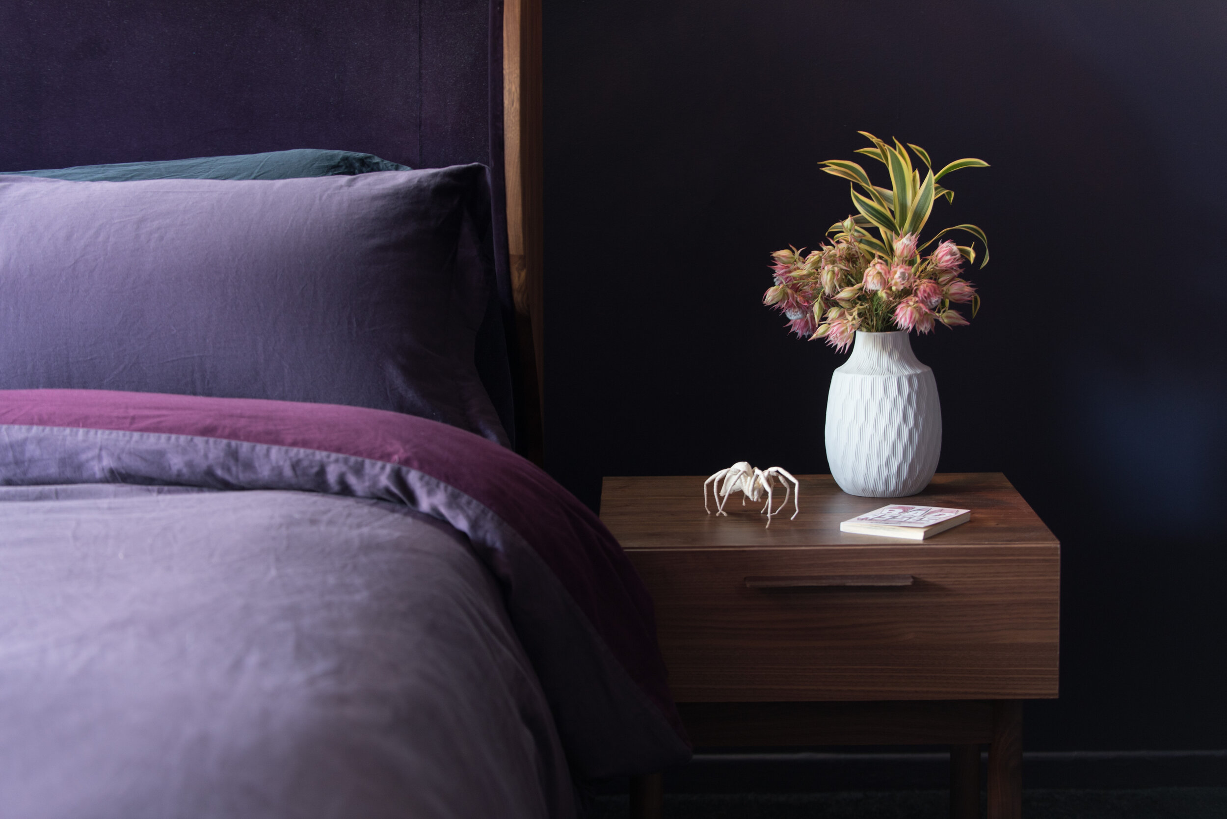Purple bed against darker purple wall, wooden side table with white vase and pink flowers, and white spider sculpture