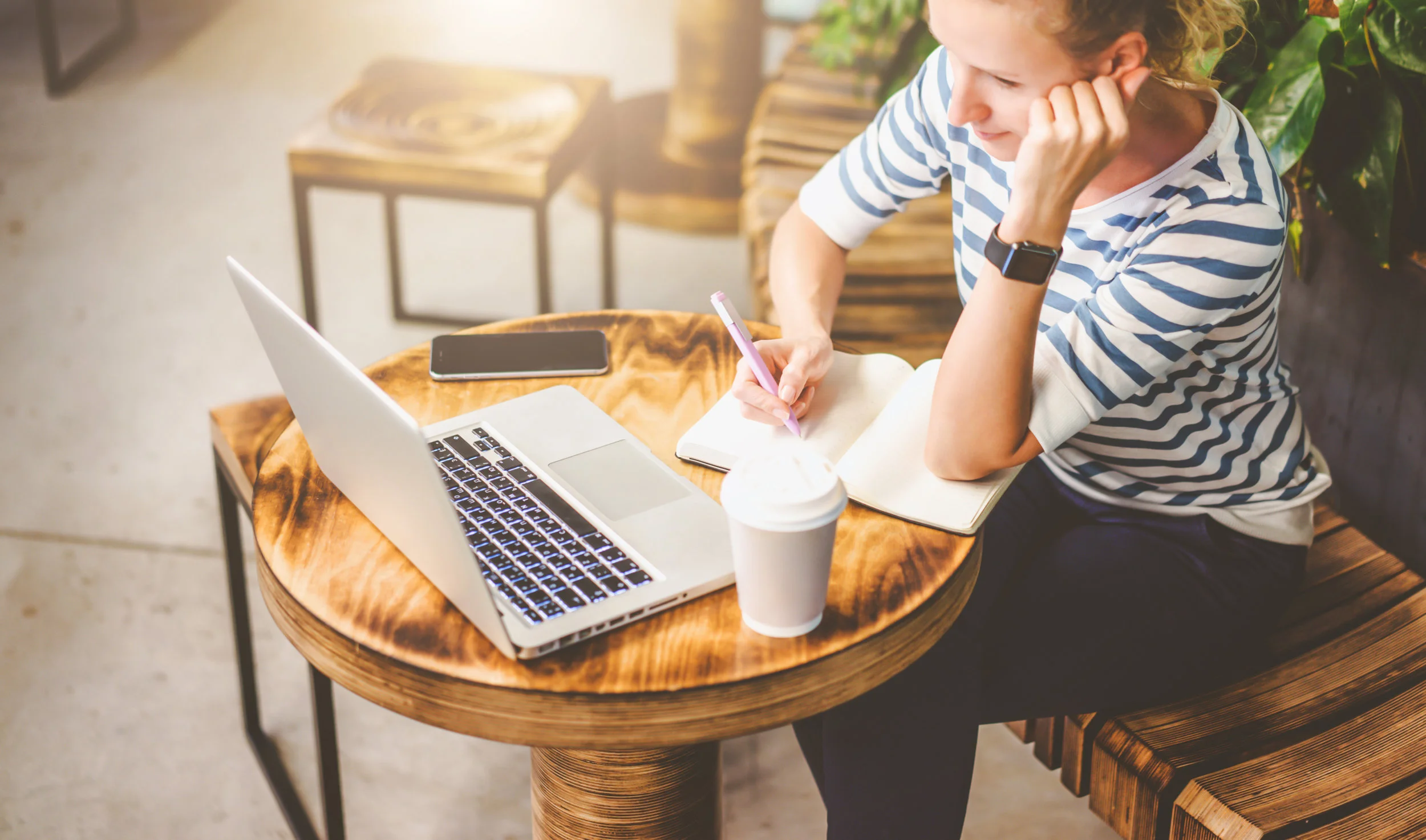View from above.Young woman in striped t-shirt sitting at round wooden table in cafe and writing in notebook.In front of her is laptop,smartphone and cup of coffee. Online learning, freelancer.