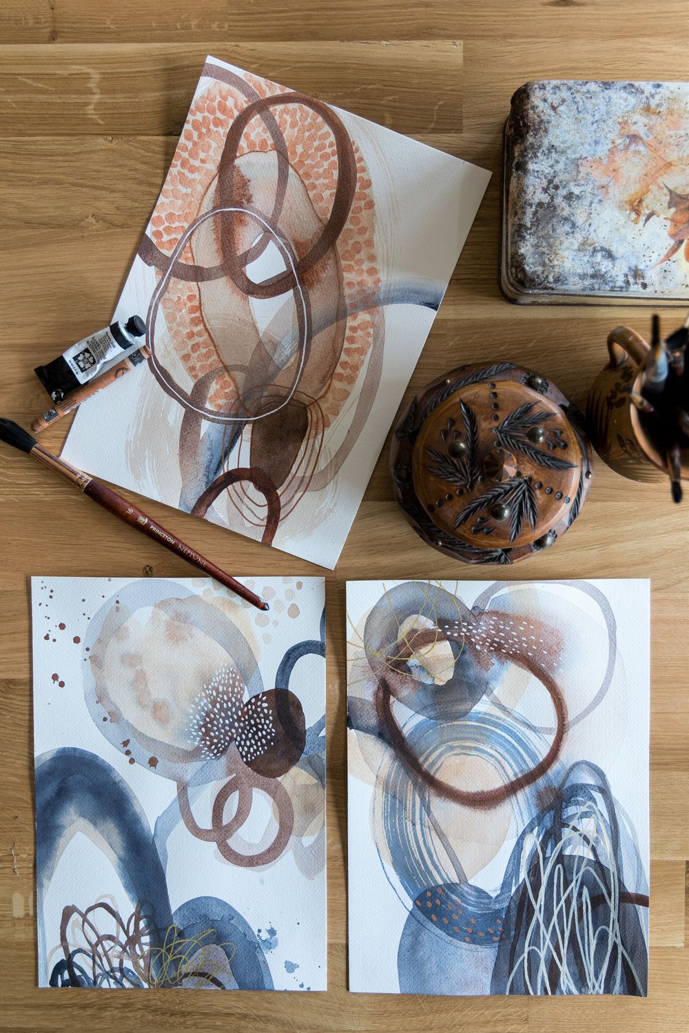  Bird’s eye view of three abstract paintings and some art supplies on a wooden table.  