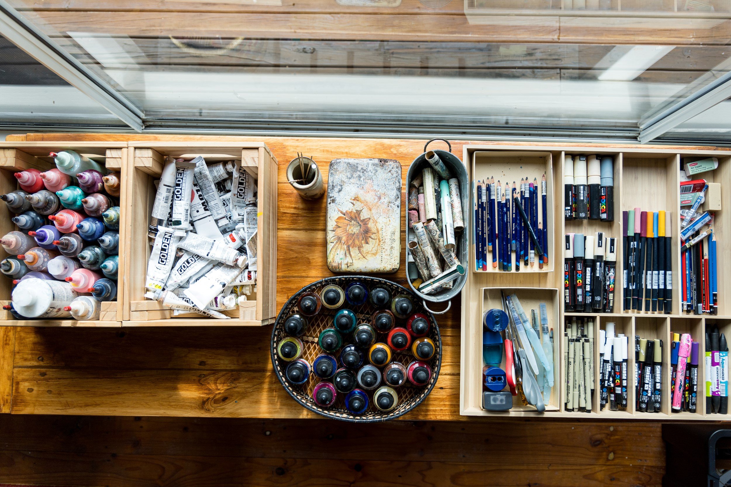  Bird’s eye view of a wooden bench with various art supplies on it, including paints, pens, inks, pastels, and pencils. 