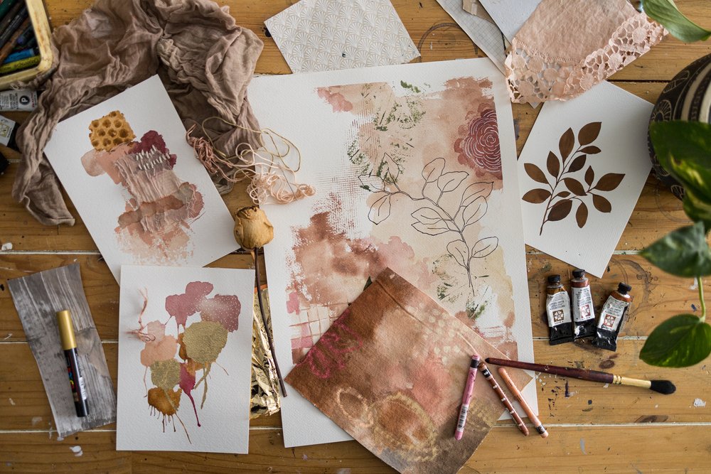  Bird’s eye view of a a wooden table with a vast array of botanical and abstract art on it as well as art supplies. A showcase of the work created in the Modern Mixed Media Course with Laura Horn.  