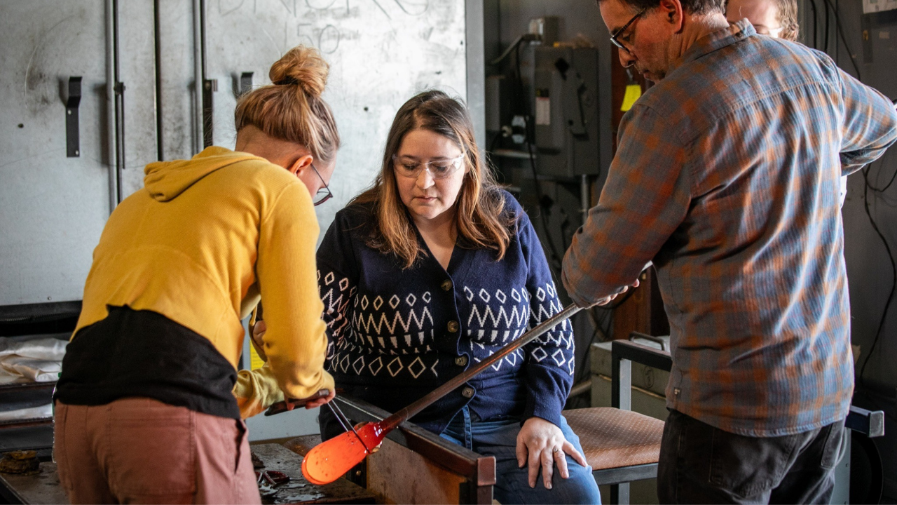 Three people working on glassblowing in a workshop, with one person shaping hot glass while the others assist, surrounded by workshop tools and equipment.