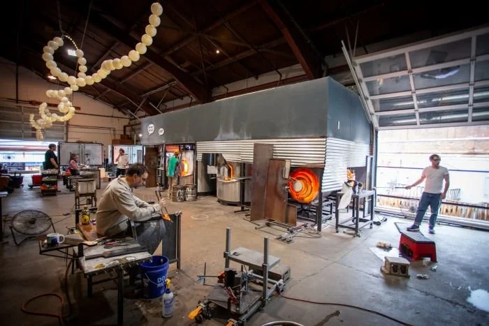 A glassblowing studio with multiple furnaces and glass art projects in progress, with people working on glassblowing and tools around.