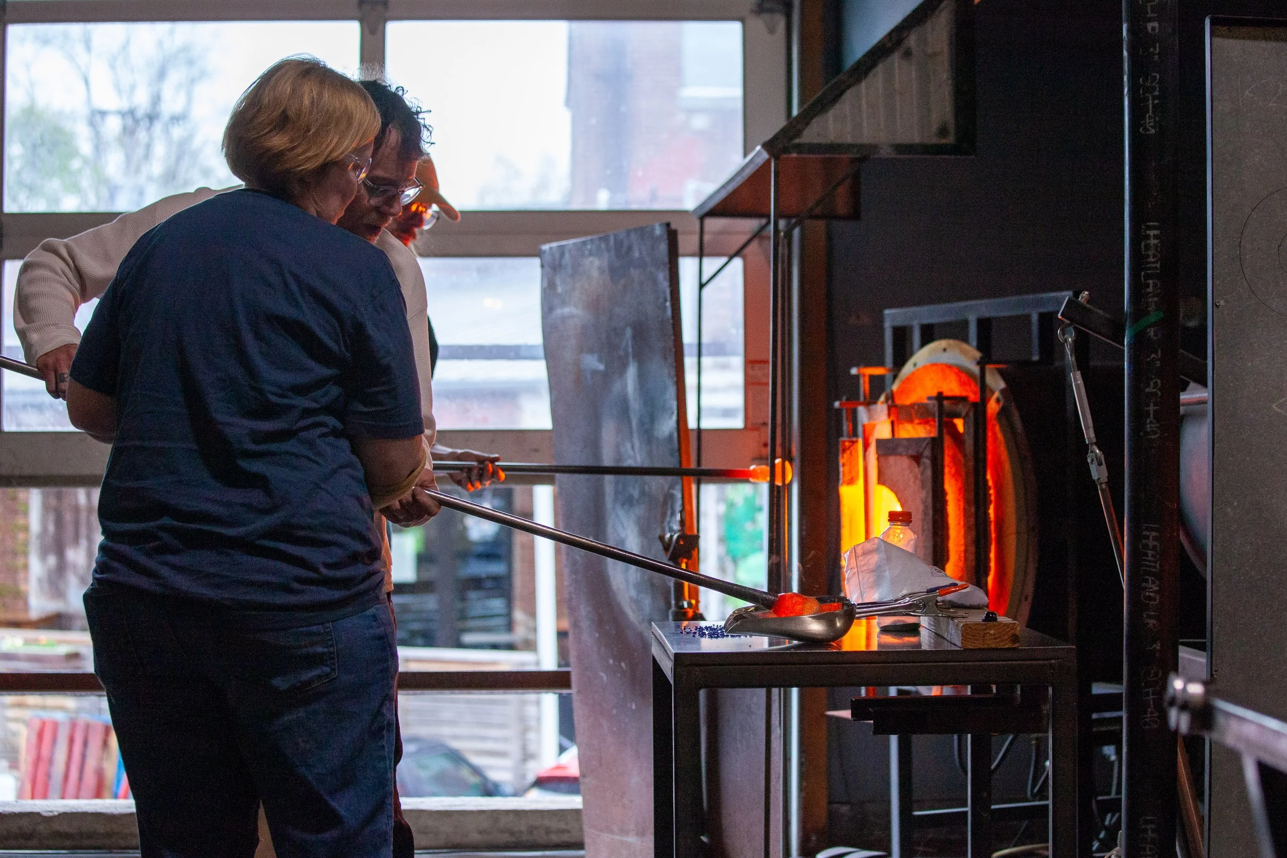 Two people coloring a glass bowl  using traditional glassblowing process at a glass studio, with molten glass glowing orange inside a furnace.