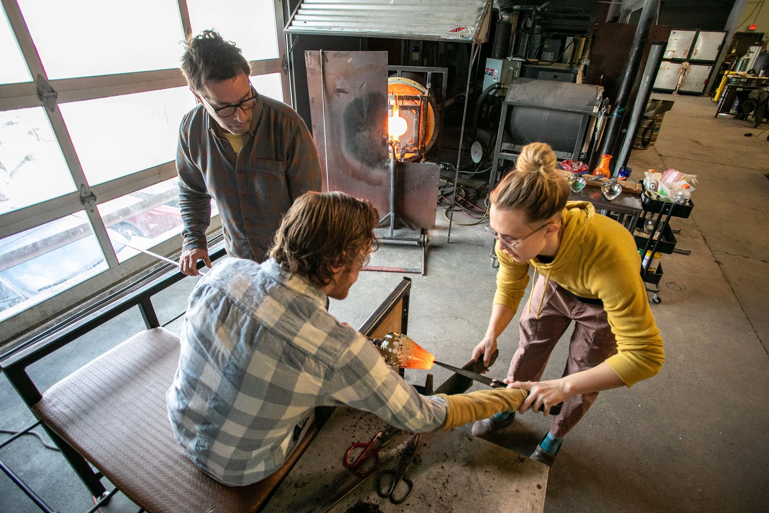 Three people working together on a glassblowing project in a workshop. A person with glasses and a striped shirt is sitting, and two others are standing, helping to shape a hot, glowing piece of glass heated in a furnace. The workshop has large windows, workbenches, and various tools and glass items on shelves.
