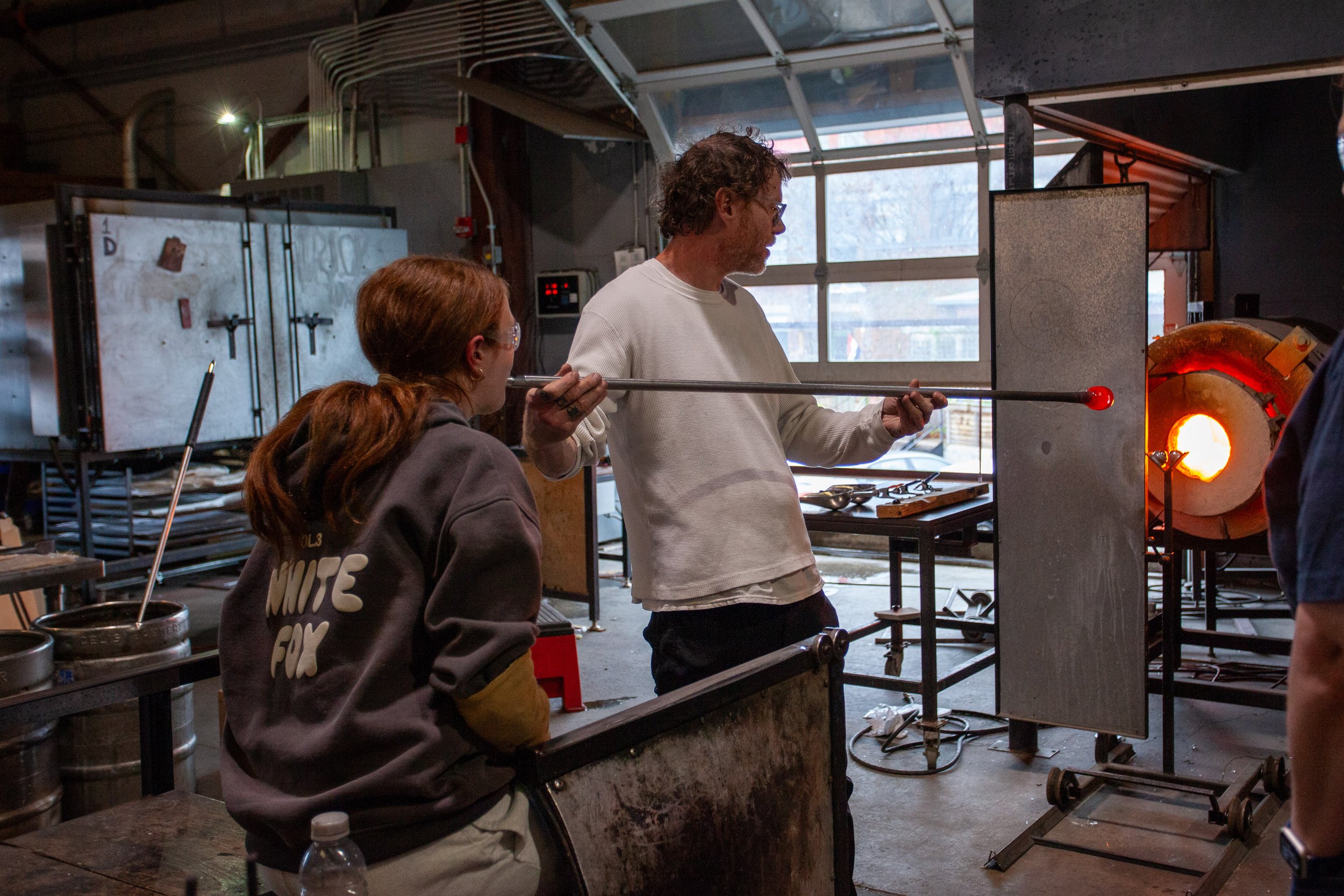 People watching a glassblowing process in a glass blowing studio with a person shaping hot glass in a furnace while others observe.