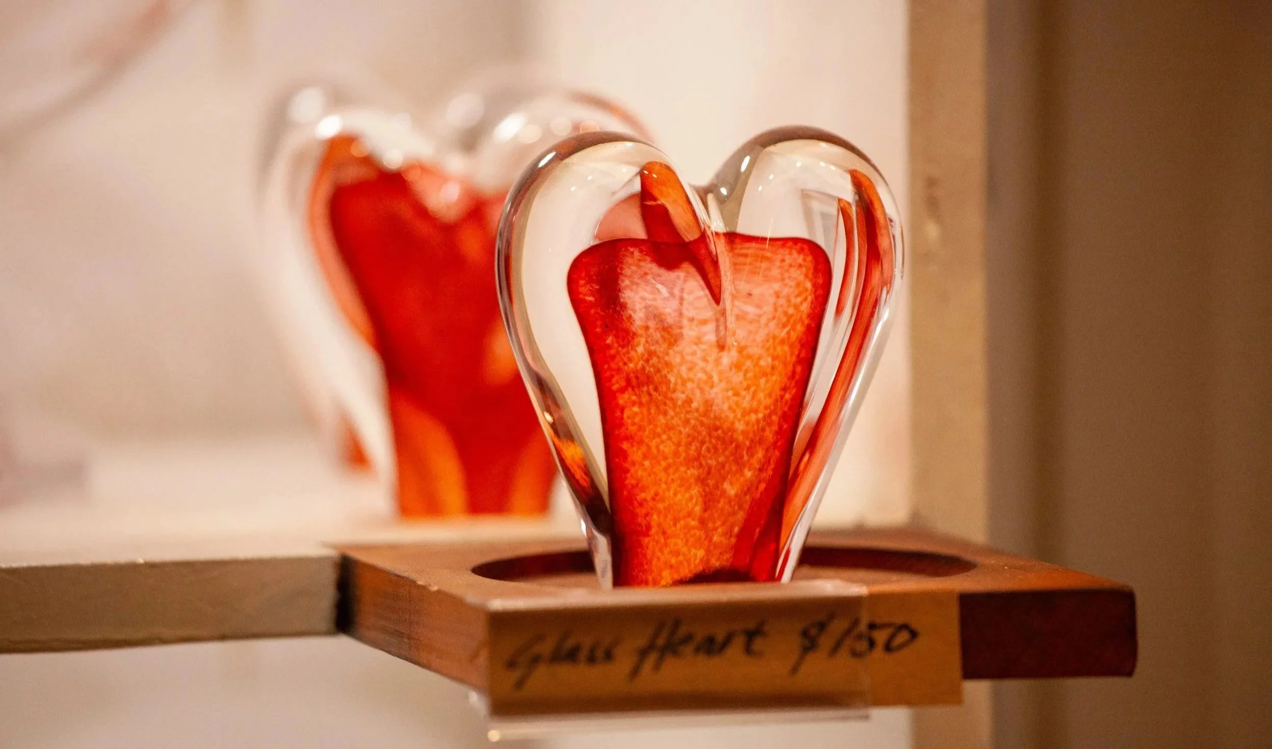 A glass heart-shaped sculpture with red and orange colors on a wooden display stand.