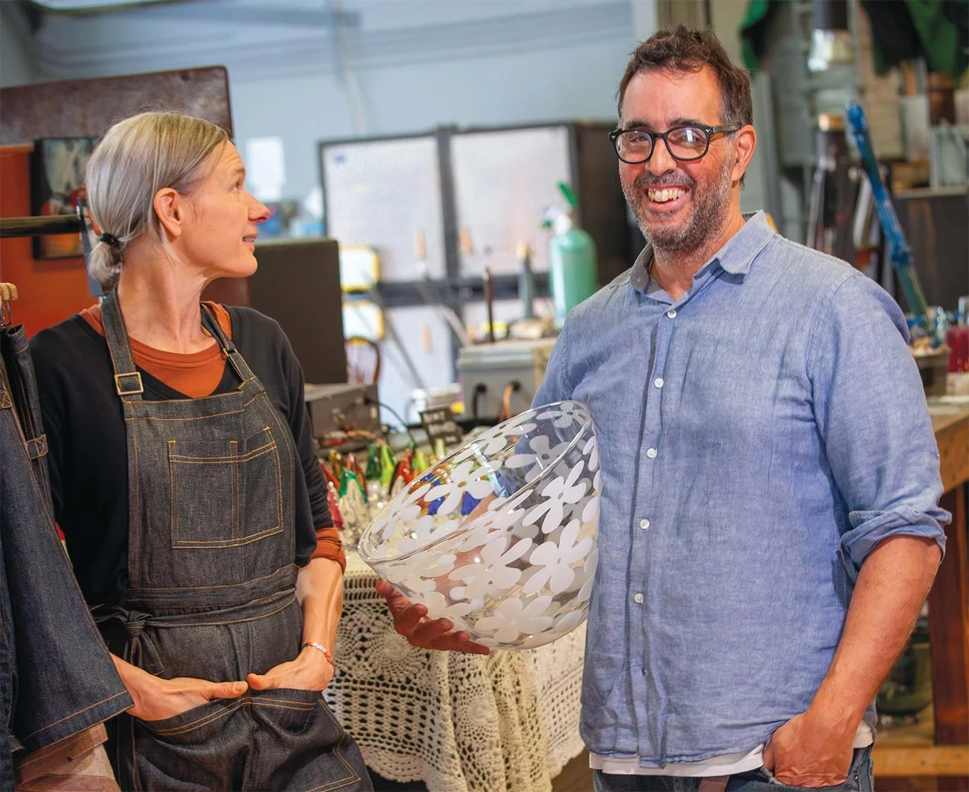 A woman in a black apron and an older man in a blue shirt smiling and holding a large flower-patterned glass bowl inside a workshop or craft store.