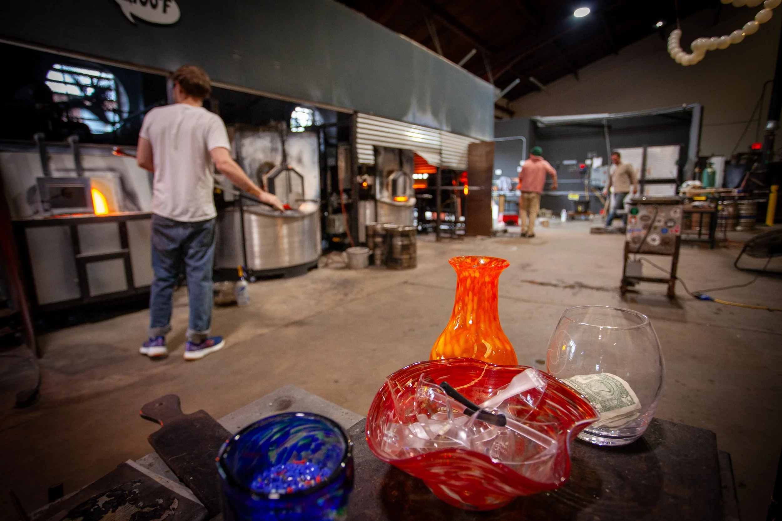 Interior of a glassblowing workshop with colorful glass pieces on the table, people working in the background, and a view of the glassmaking process.