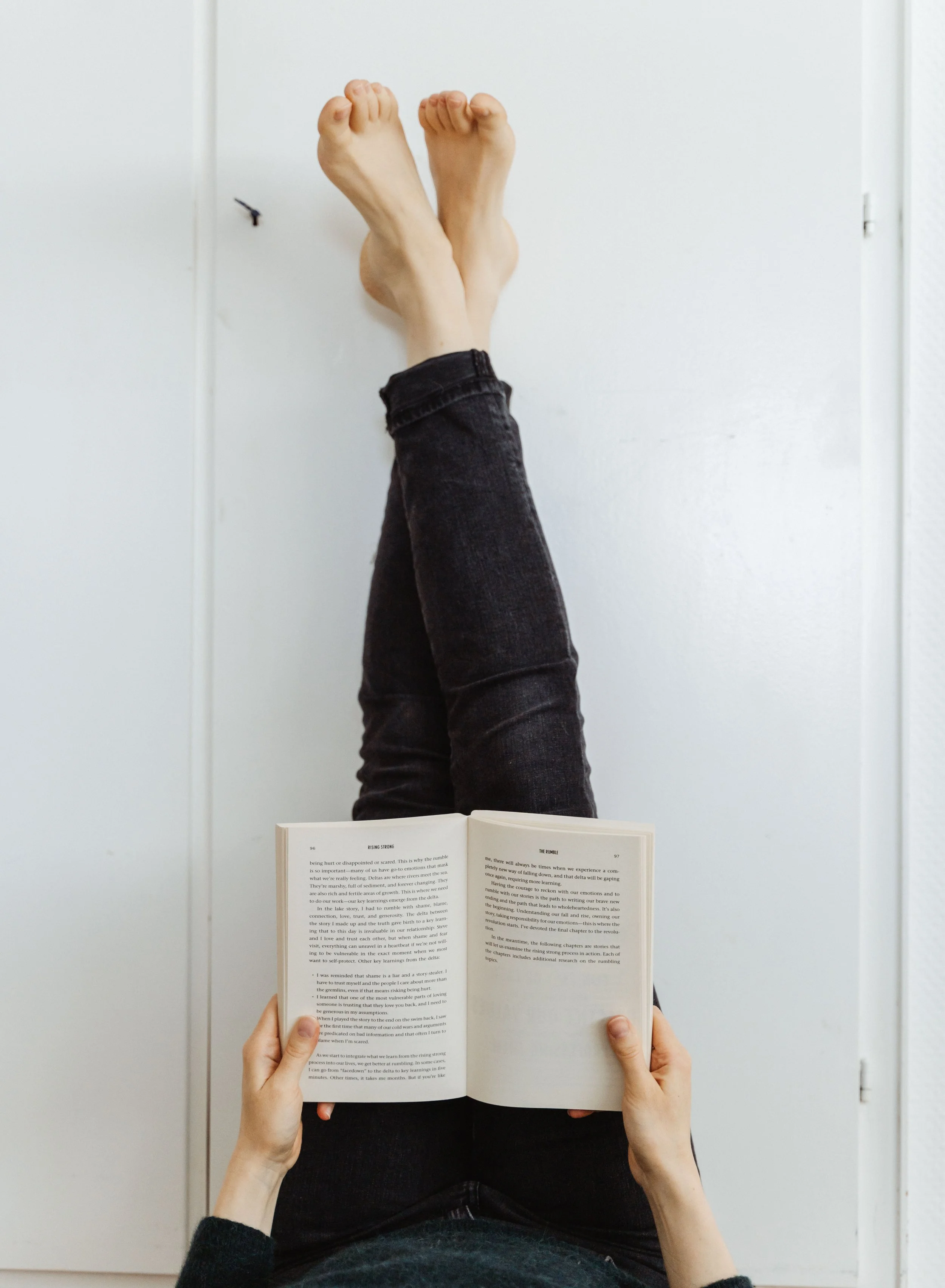 Person reading a book with feet propped against a white wall, wearing dark jeans.