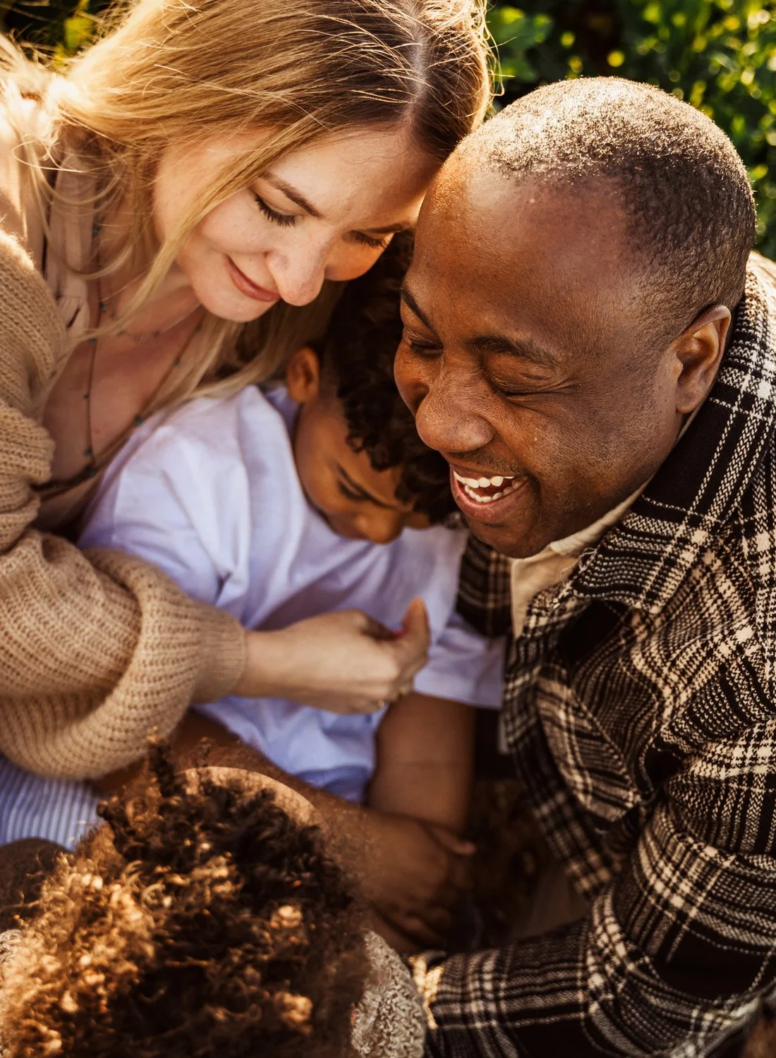 A joyful family scene depicting two adults and two children, gathered closely together, sharing a warm hug outdoors with sunlit greenery in the background.