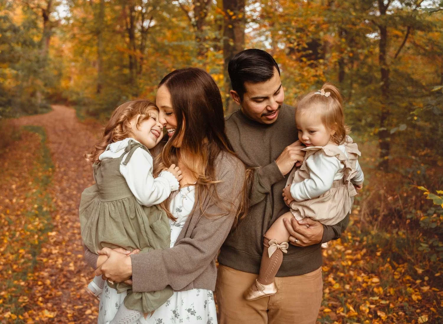 young family of four with two young daughters. parents are holding girls up and close and snuggle in beautiful fall scenery on a sunny day in fall in germany. they are all smiling and laughing at each other