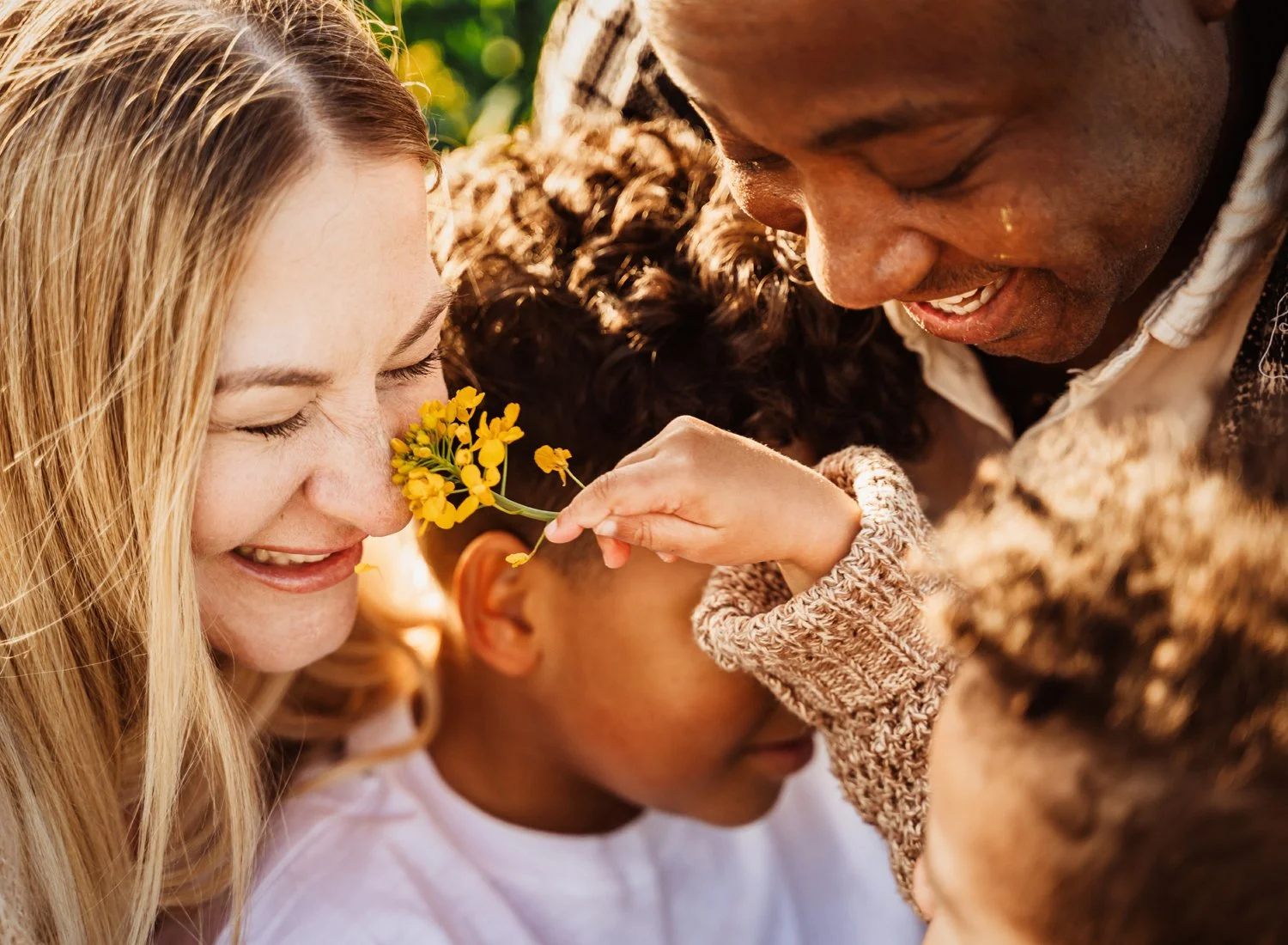 A close-up of a smiling family enjoying time together outside. A child holds a yellow flower near an adult's face.