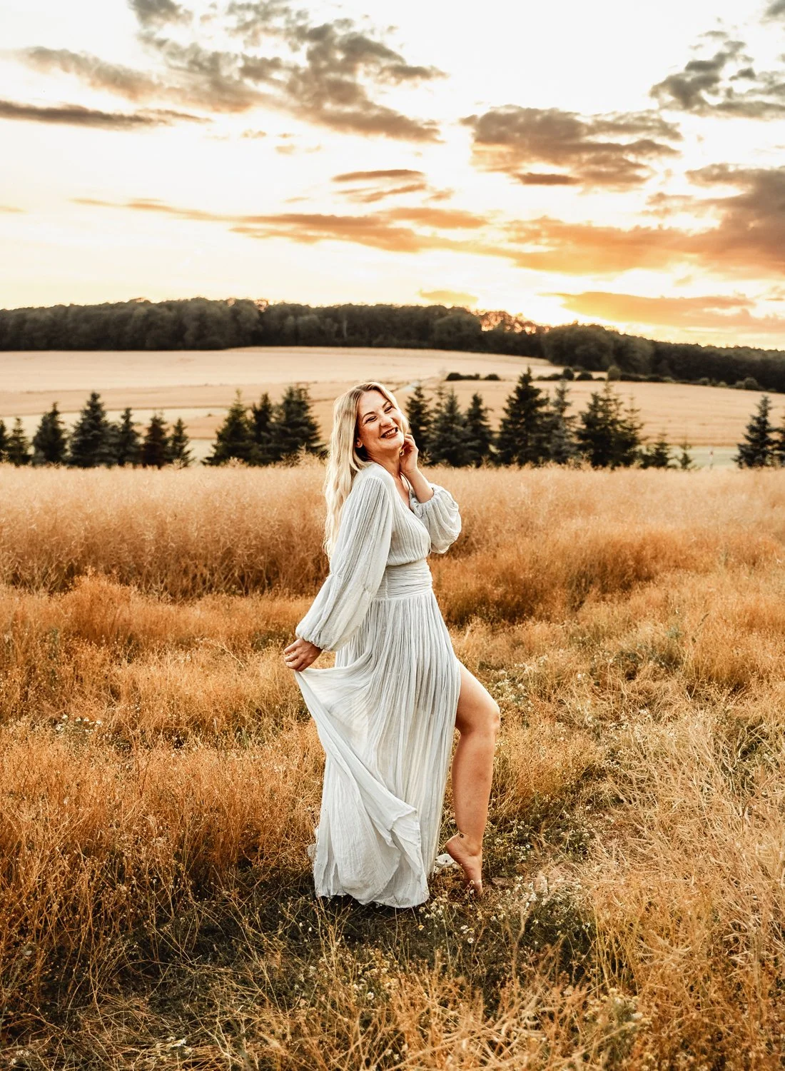 Woman in white dress smiling in a field with a sunset and pine trees in the background.