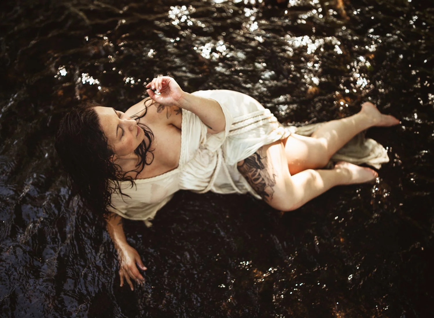 Woman in white dress lying on rocks in a stream, sunlight reflecting on water.