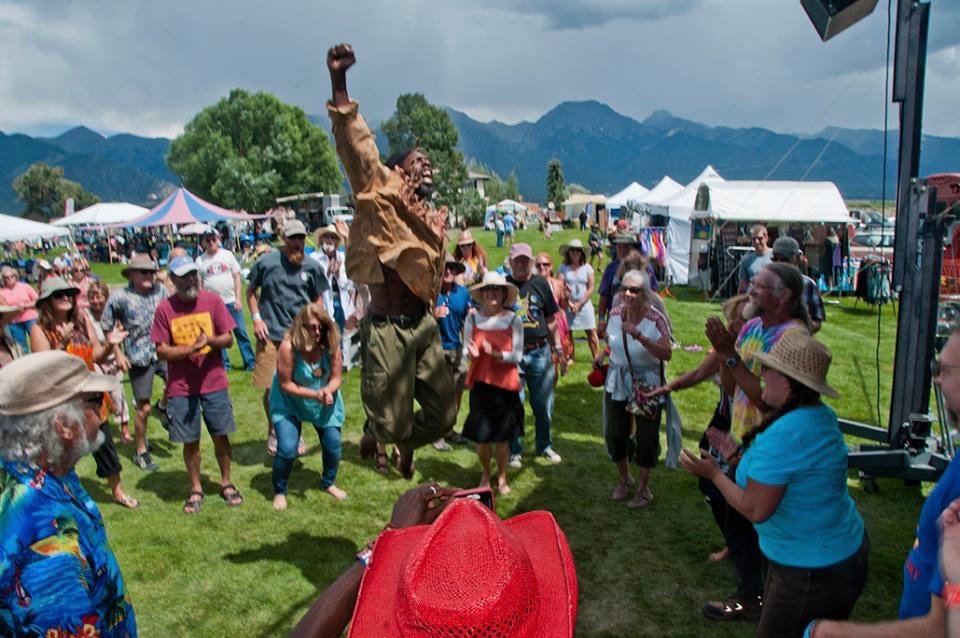An airborne performer raises a fist beside the stage, surrounded by festivalgoers, with tents and mountains behind.