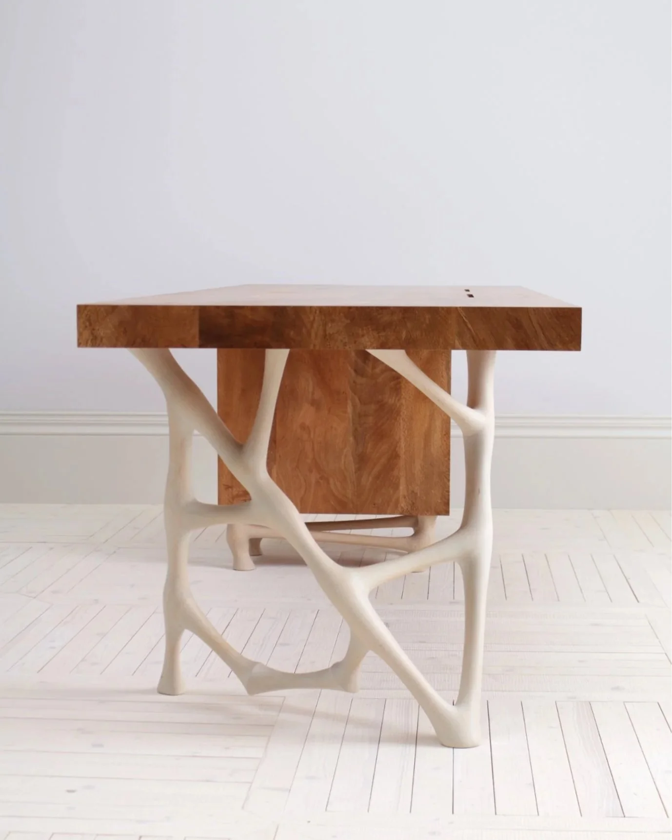 A wooden table with a top and base made of natural-colored wood, supported by sculptural, white, branch-like legs, set against a plain white wall with a white floor.