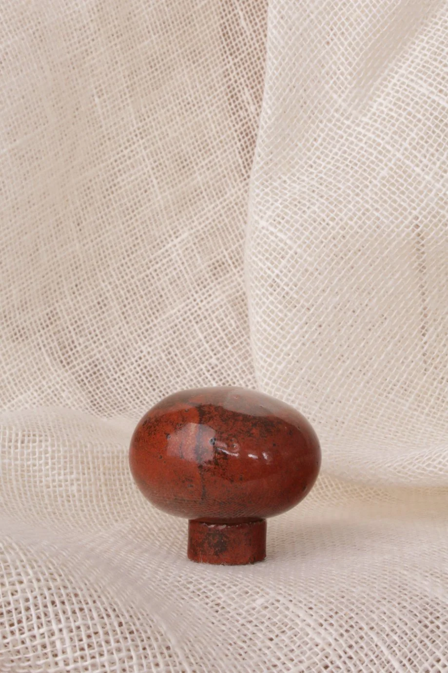 A smooth, polished, reddish-brown stone with black and dark red markings, resting on a small round base, against a textured, cream-colored fabric background.