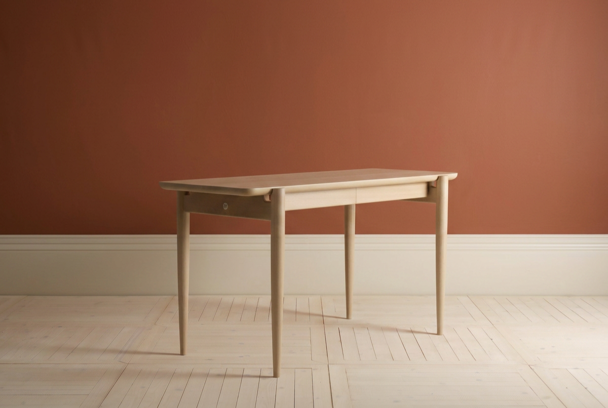 A light wooden desk with tapered legs against a pinkish wall, on a light-colored wooden floor.
