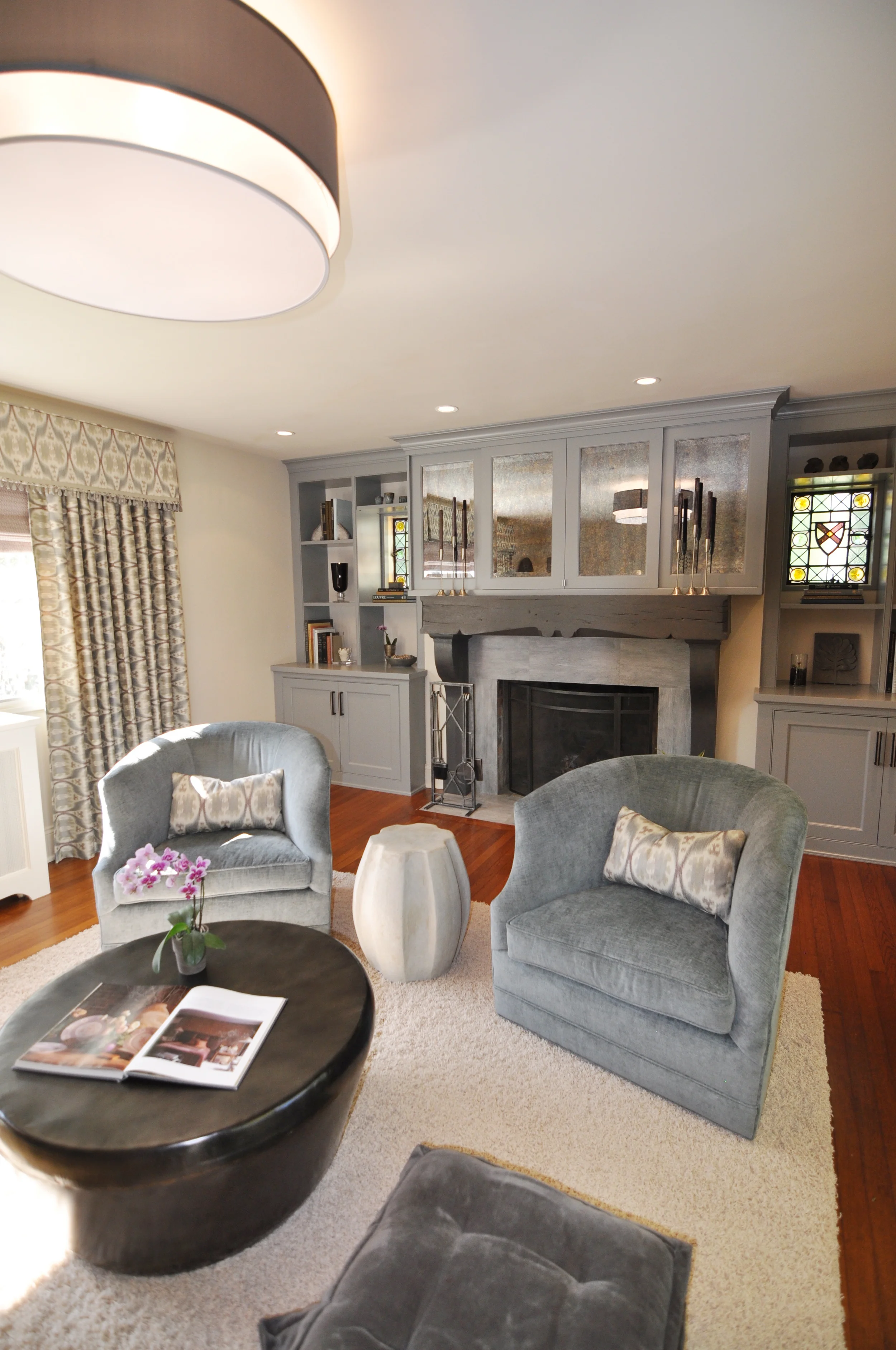 TRANSITIONAL BOHEMIAN LIVING ROOM , Larchmont, NY: A 1920’s Tudor home receives a contemporary makeover to create an inviting living space fit for adult and family time. Sail cloth colored paint on re-plastered walls smooths cracked and uneven walls. A blue-gray fabric double drum shade fixture establishes the main seating area. New ceiling lights highlight the built-in and fireplace facade of large subtle metallic porcelain tiles. Custom-designed built-in showcases the original stained glass windows and includes storage for books and toys. As this living room is also the family room, blue and plum antique mirrored sliding panel doors hide the TV when a more formal look is desired. Contemporary, yet family-friendly furnishings include swivel chairs, a low metal drum cocktail table and floor pillows. Ikat patterned window treatments accented with a wood-bead trimmed valance infuse a touch of bohemian flair, completing the chic, yet relaxed family space.
