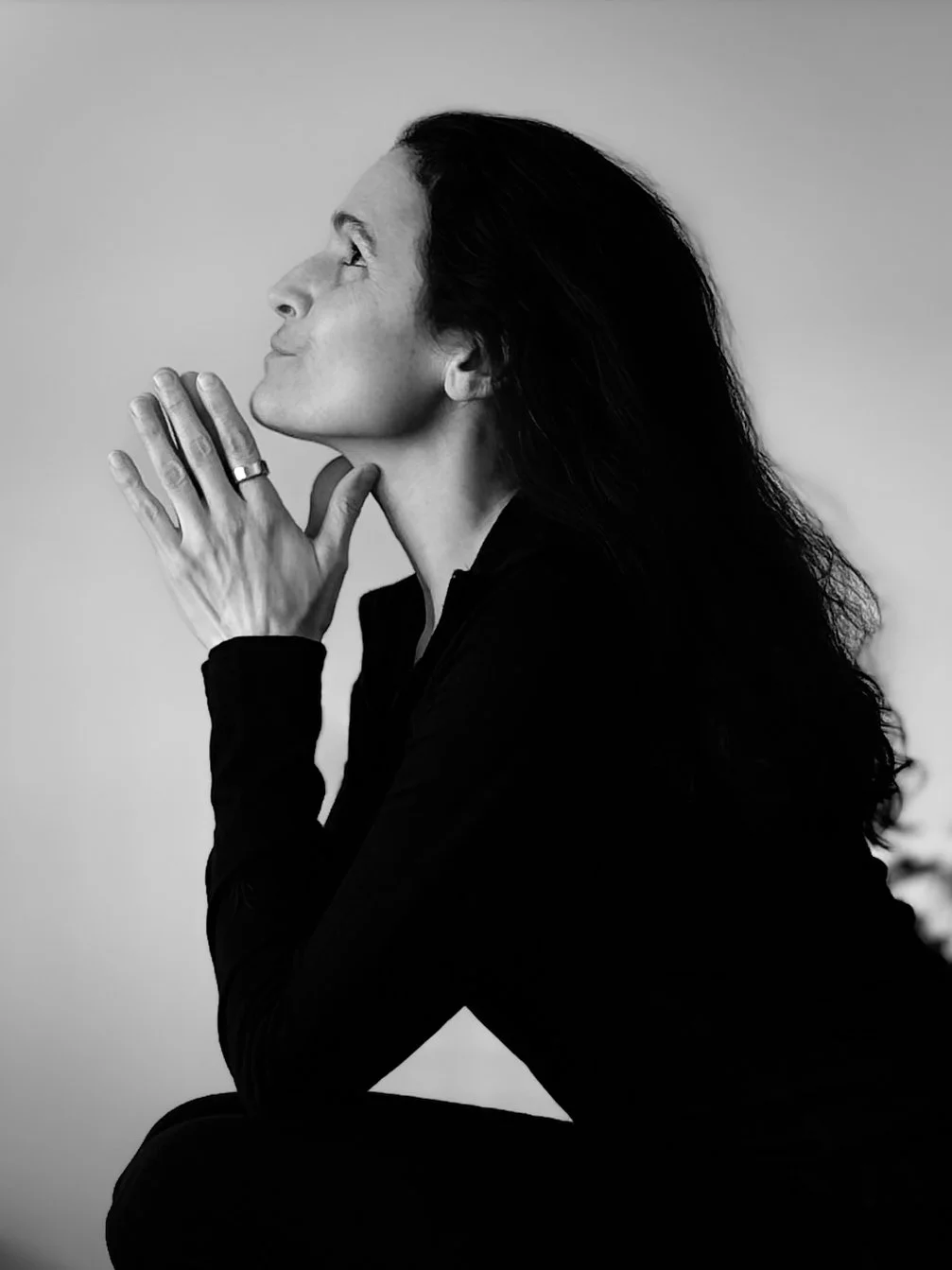 A woman with long dark hair sits in profile, her hands clasped near her chin, eyes closed, smiling gently.