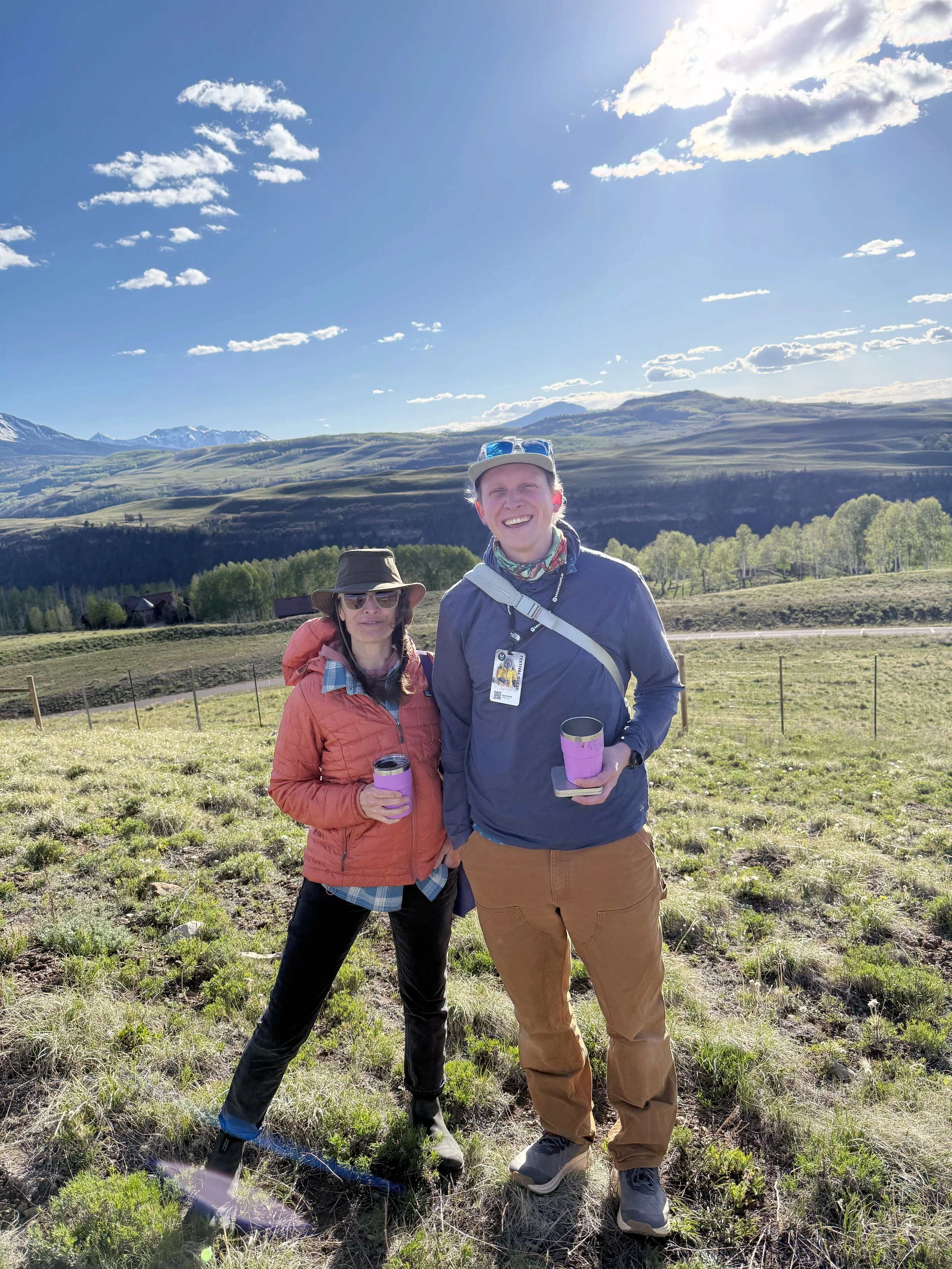 Two people standing outdoors in a grassy field with mountains and a blue sky with clouds in the background. They are smiling and holding cups, dressed in outdoor hiking clothing.