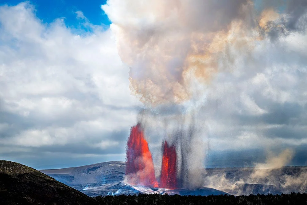 Daytime volcanic eruption with twin lava fountains and smoke rising into a lightly clouded sky at Kīlauea Hawaii.