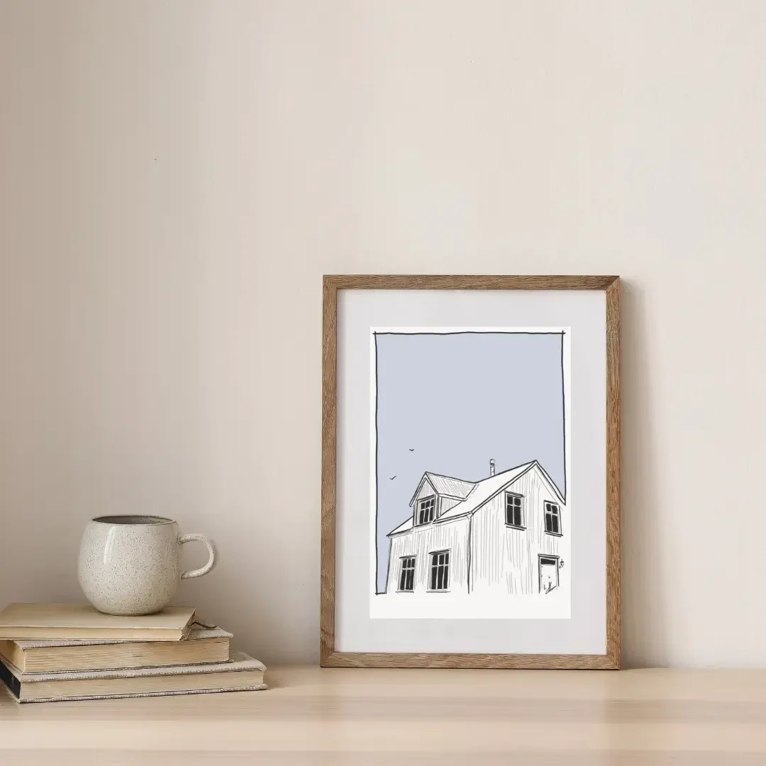 Framed digital line drawing of a traditional white Icelandic house in Reykjavík, styled on a wooden shelf with stacked books and a ceramic mug in a calm Scandinavian interior.