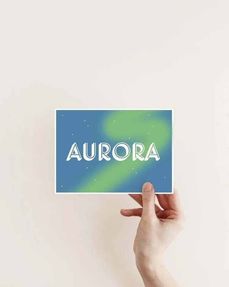 Hand holding a postcard featuring the word “Aurora” over a stylised Northern Lights sky in blue and green, minimalist Iceland-inspired typography art.
