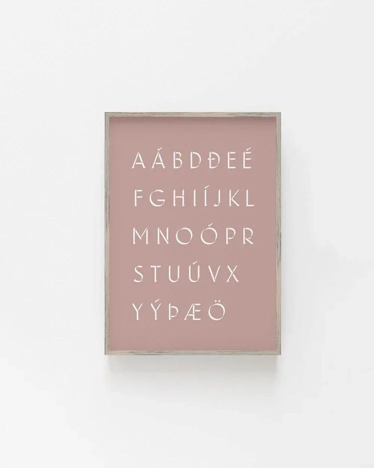 Framed Icelandic alphabet typography print in white lettering on a soft pink background, minimalist Scandinavian wall art for nursery or modern home decor.