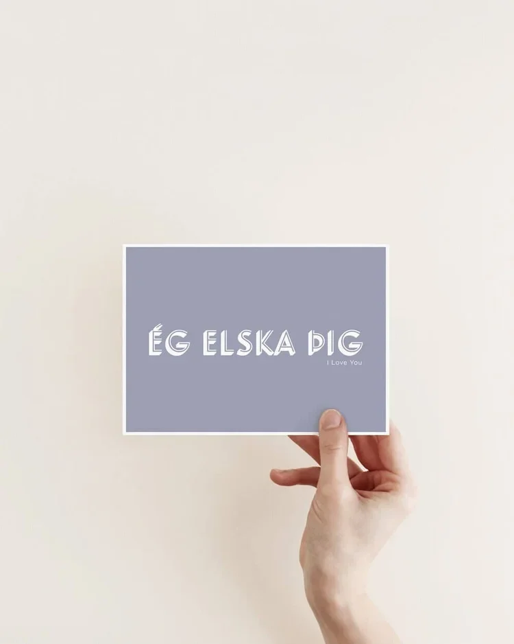 Hand holding a minimalist postcard with the Icelandic phrase “Ég elska þig” meaning I love you, in white typography on a soft blue background.