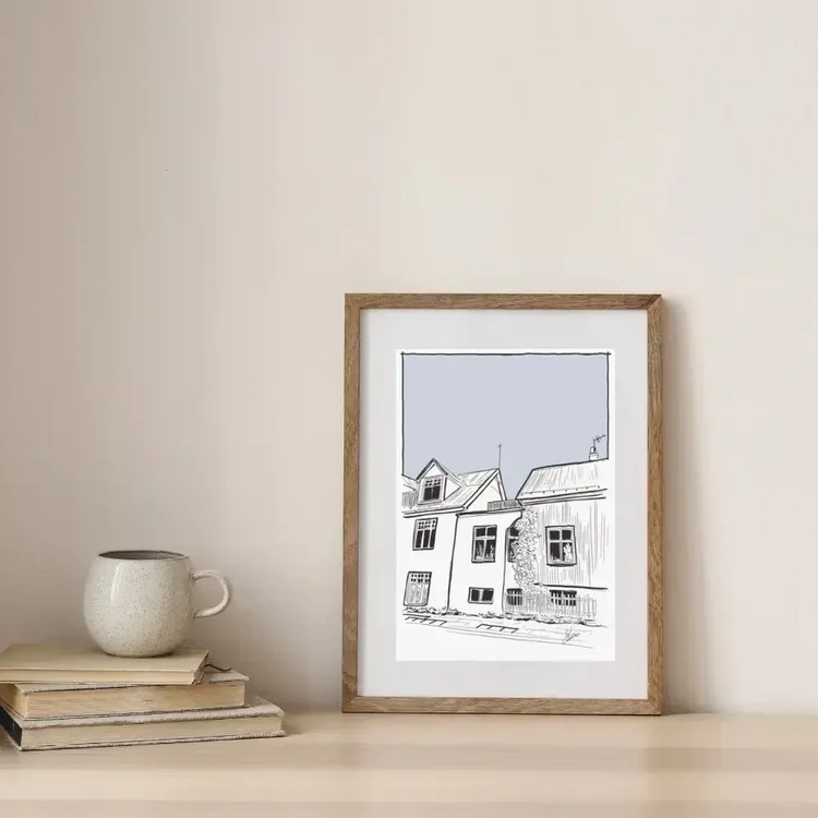 Framed digital architectural line drawing of traditional houses on Spítalastígur in Reykjavík, Iceland, styled on a wooden shelf with books and a ceramic mug in a neutral Nordic interior.