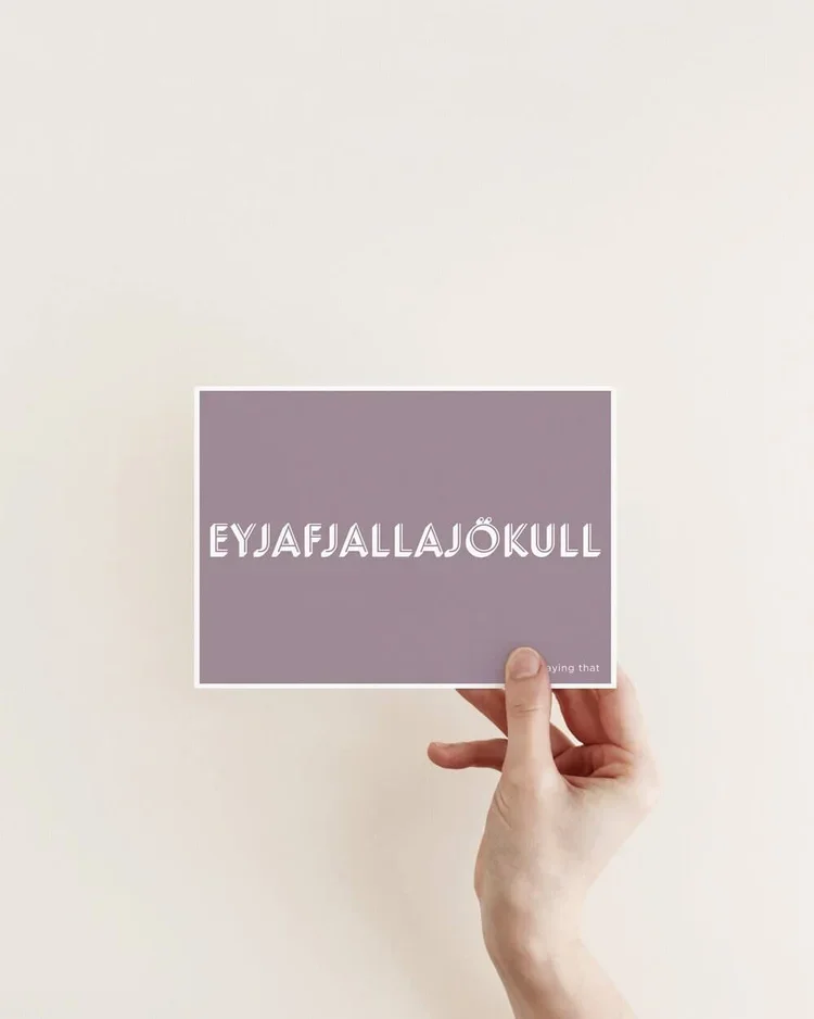 Hand holding a minimalist postcard with the Icelandic word “Eyjafjallajökull” in white typography on a muted purple background.