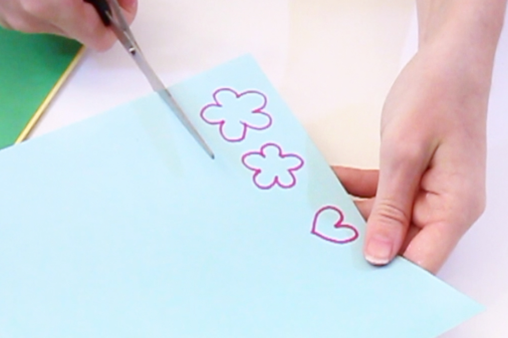 Step 6 Draw the shapes of two flowers and a heart onto the blue piece of card and cut out. Then cut two circles out of the yellow card (for the inner bit of the flower) and two stem shapes out of the green card. Glue the yellow circle onto the blue flower and the flower onto the stem.