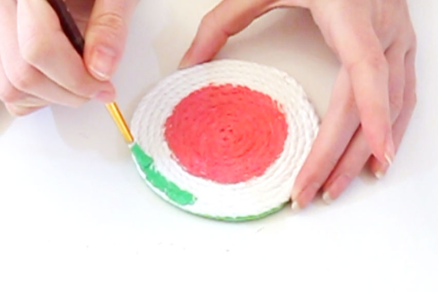 Step 3 Mix the red and white paint together until you have the colour for the centre of the watermelon. Paint a circle in the middle of the coaster with this paint. Then paint around the edge of the rope with the green paint, leaving a small white line in-between the two colours. Leave to dry. Once the red paint is dry, paint some small black dots over the top of it to create the watermelon seeds.