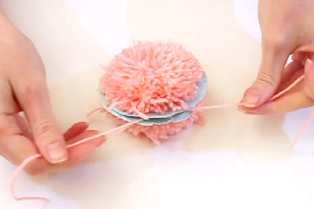 Step 3 Tie a piece of wool of the same colour around the pom pom in between the two pieces of card. Make sure you pull it tightly and tie in a double knot! Trim the ends of this wool and rip off the card and you have your pom pom.