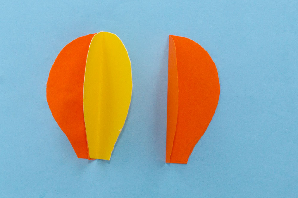 Step 2 Fold all of the hot air balloon shapes in half. Then, take one piece and glue one half to the blue paper. Glue the other half and stick the next piece to it, as shown above.
