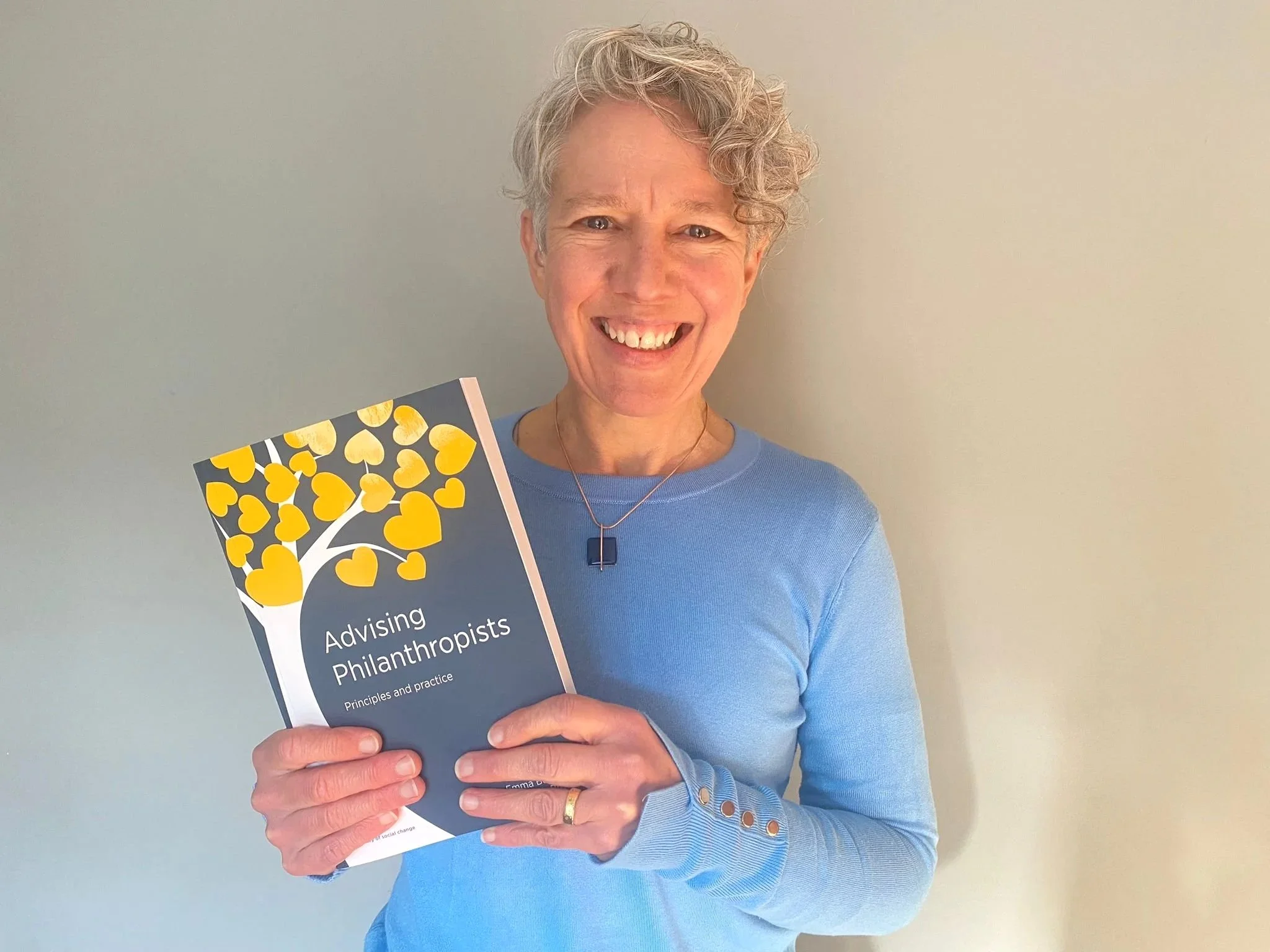 A smiling woman with short wavy grey hair wearing a blue jumper, holding a book titled "Advising Philanthropists: Principles and Practice" in front of a plain light-coloured wall.