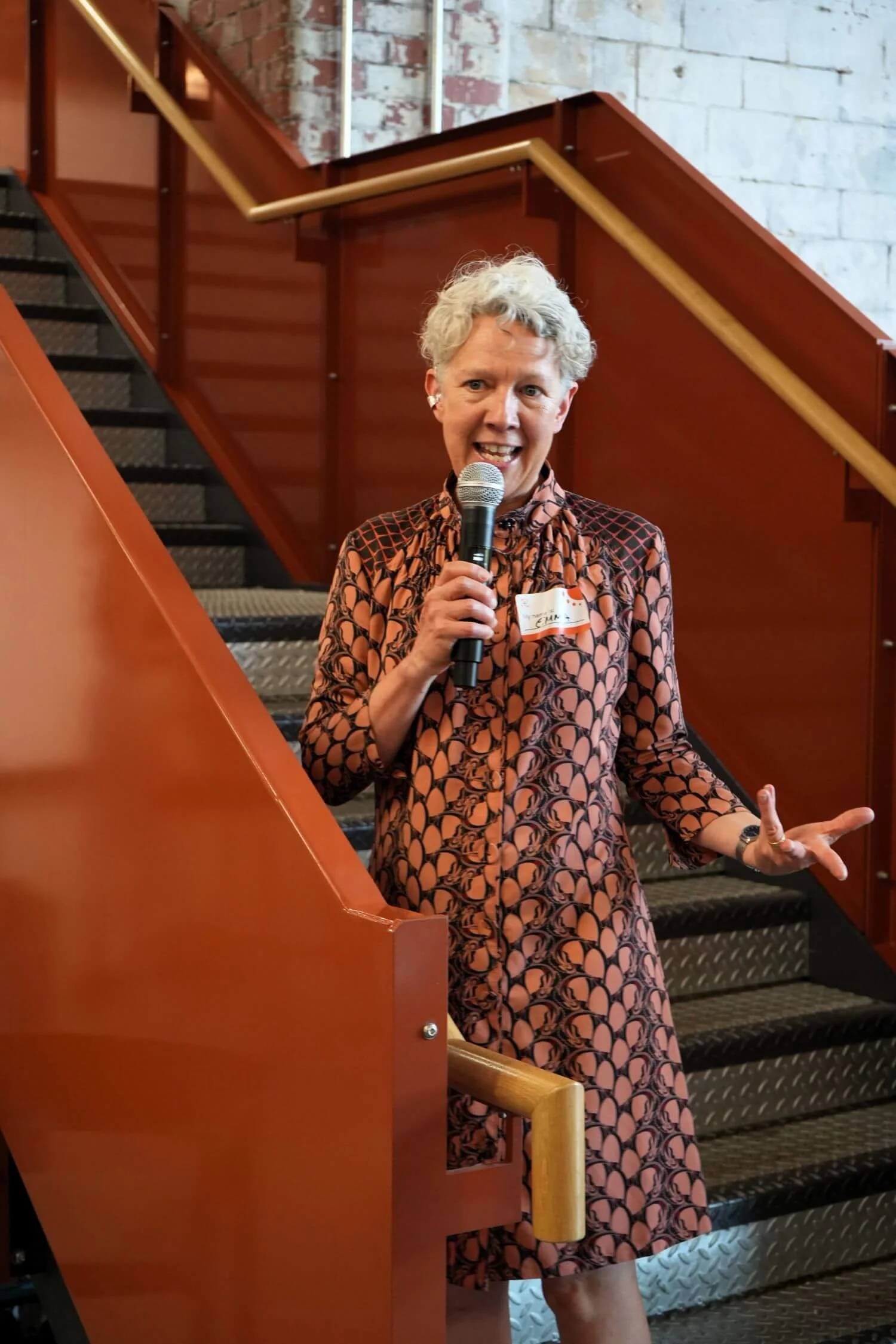 A woman with short wavy grae hair holding a microphone and gesturing with her other hand, standing on a staircase.