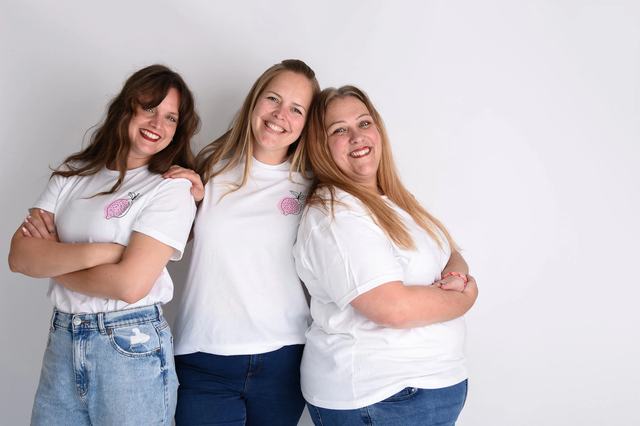 3 ladies stood in the studio smiling at the camera. They are wearing white tshirts with a pink lemonade logo and blue jeans