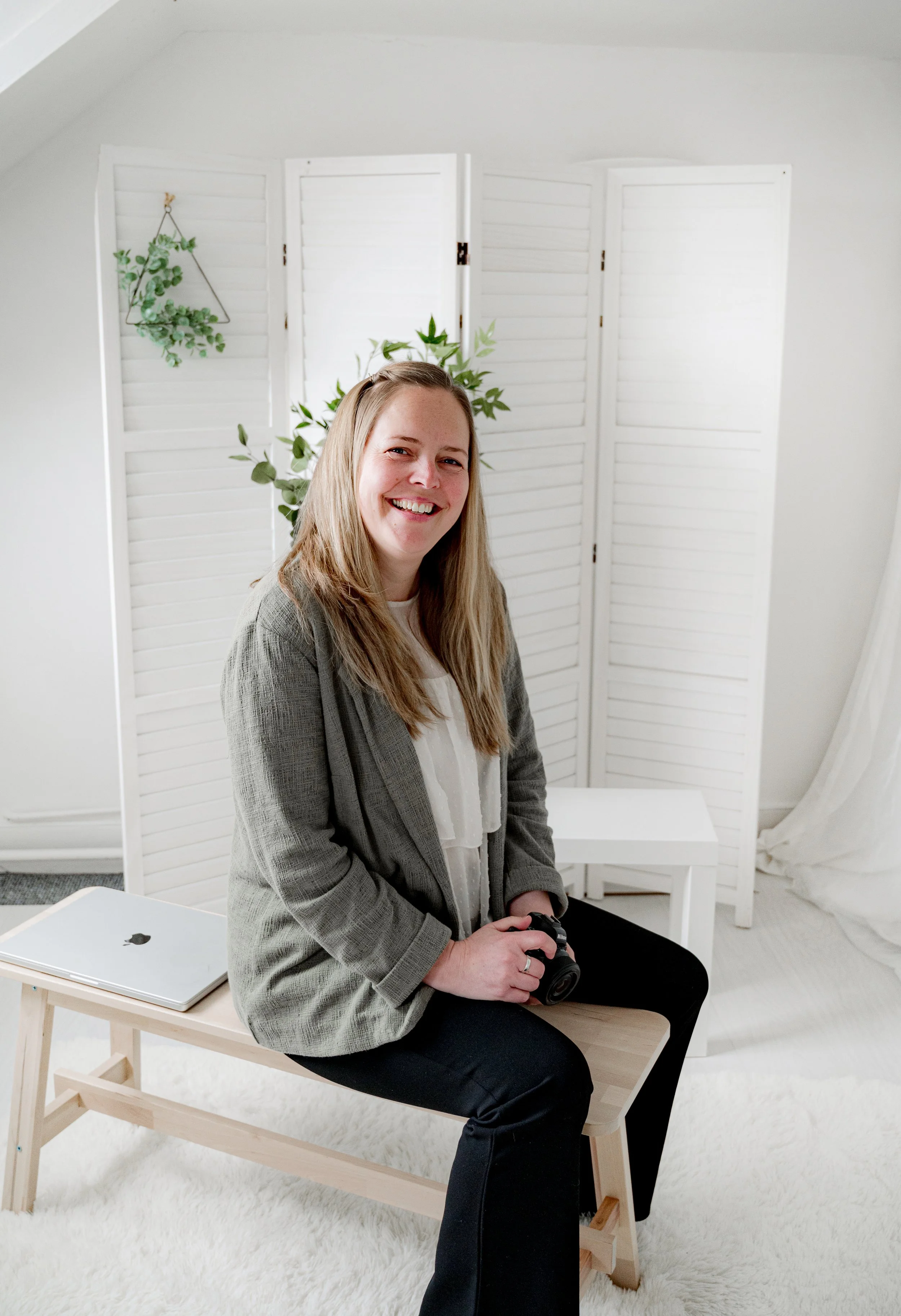 A woman with long blonde hair sitting on a wooden bench in a bright white room, smiling, with a green plant behind her, a laptop on the bench, and a folded camera in her hands.