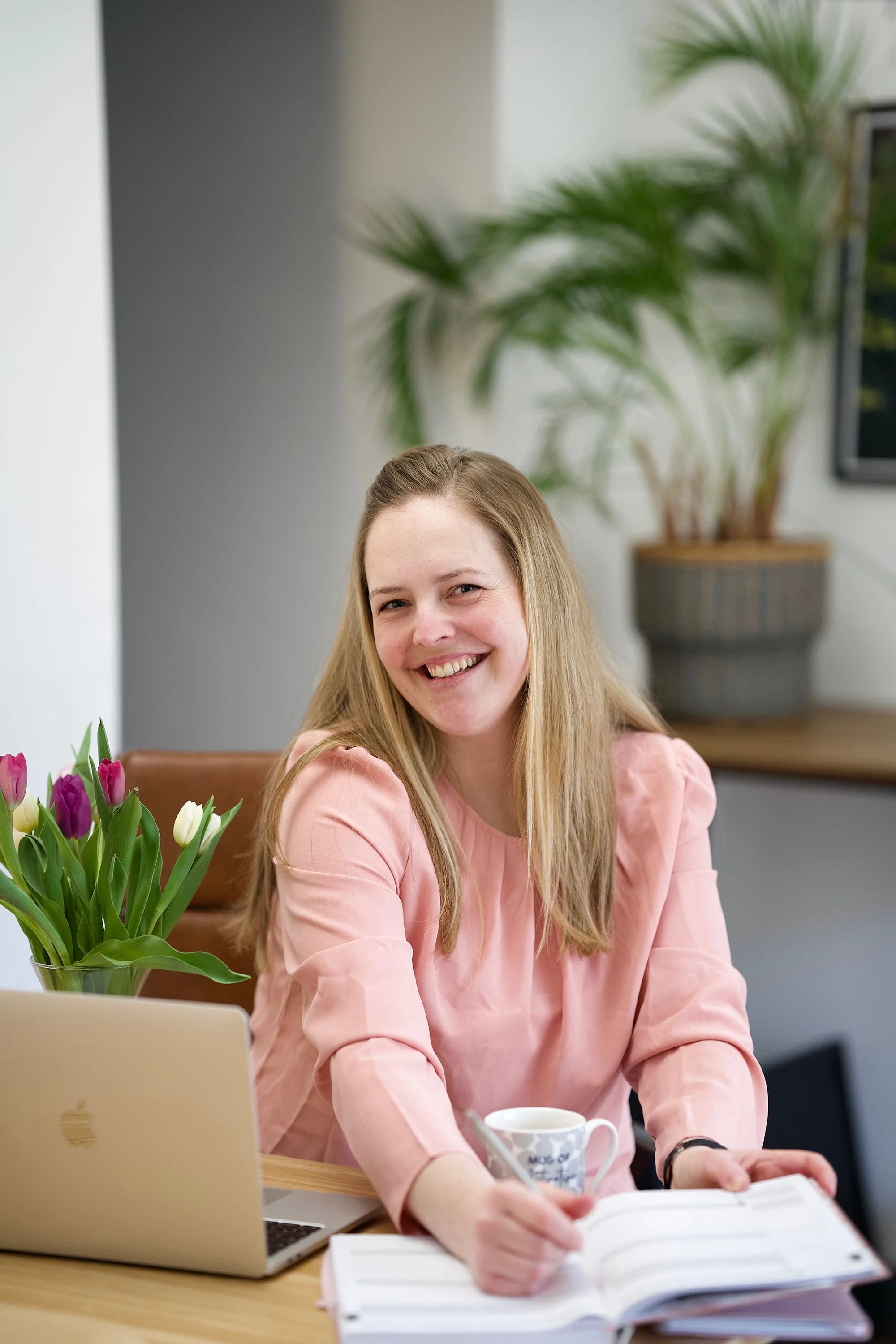 A woman sitting at a desk with an open notebook, a mug, a laptop, and a vase of tulips in the foreground, smiling at the camera. In the background, there is a large potted plant and a wall with a framed picture.