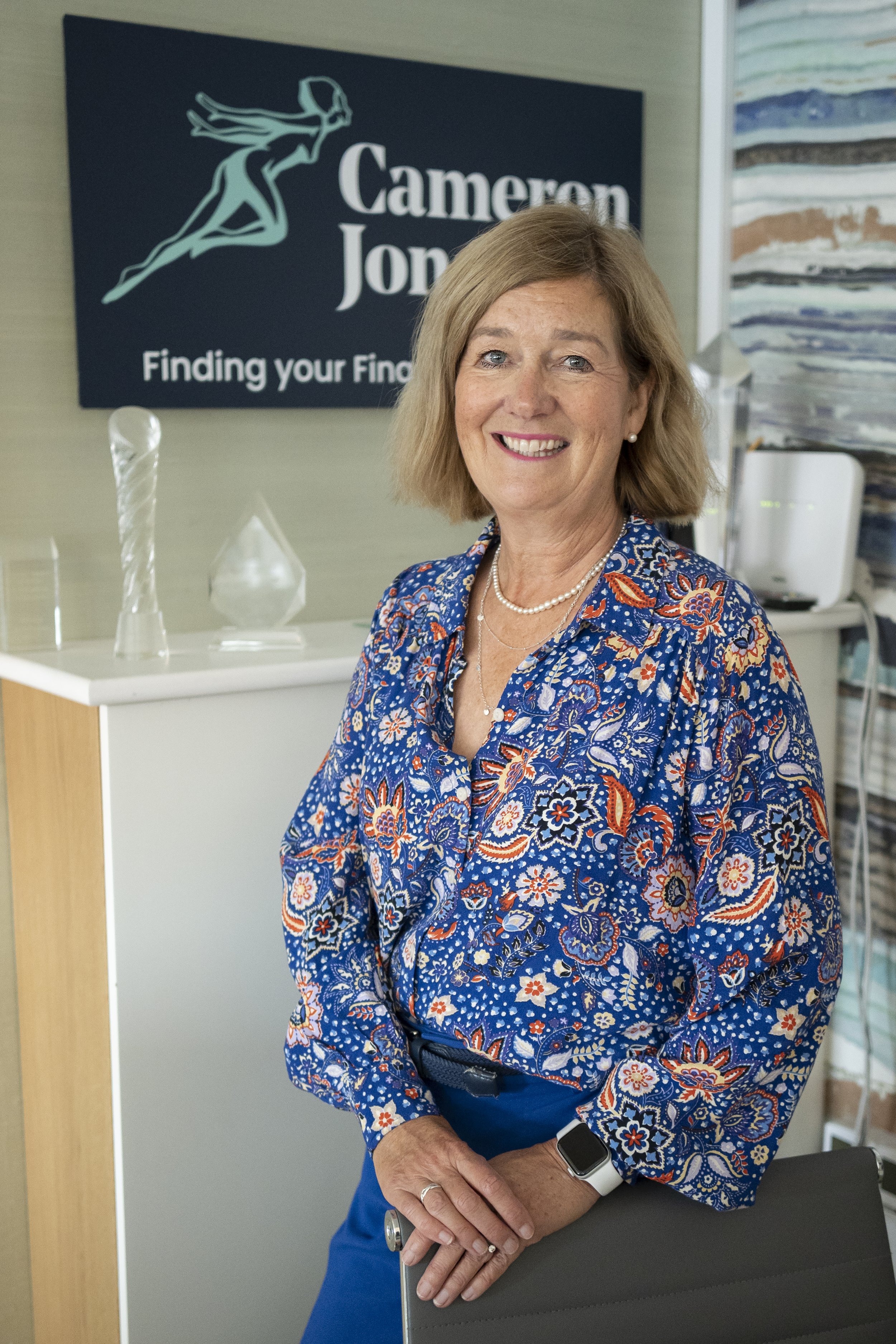 headshot of lady in her office. She is stood next to the chair wearing a blue patterned top.